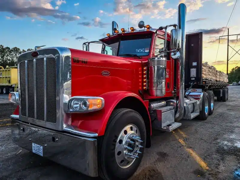 Bright red Peterbilt semi-truck with chrome details and flatbed trailer loaded with materials, parked on gravel lot at sunset.