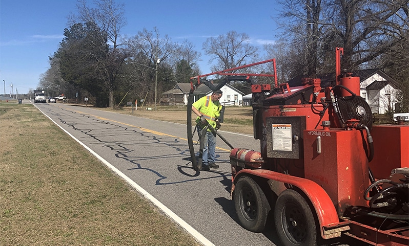 Worker applying sealant into pavement cracks using specialized crack-sealing equipment on asphalt surface